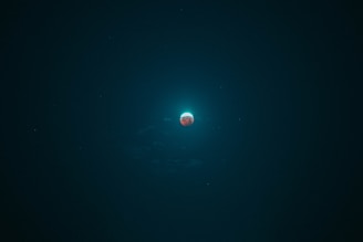 A celestial body glows with a bright halo against a dark sky. The object appears to be a planet or moon with a reddish surface, surrounded by a blue-green aura. Tiny stars or celestial bodies are faintly visible around it.