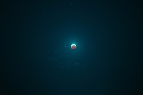 A celestial body glows with a bright halo against a dark sky. The object appears to be a planet or moon with a reddish surface, surrounded by a blue-green aura. Tiny stars or celestial bodies are faintly visible around it.