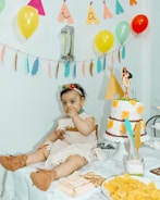 A baby dressed in a costume with a headband and brown booties sits on a table covered with a lace cloth. The table is decorated with snacks, including wafers and chips, and a cake adorned with yellow flowers and a figurine on top. Balloons and colorful decorations hang in the background, creating a festive atmosphere.