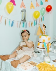 A baby dressed in a costume with a headband and brown booties sits on a table covered with a lace cloth. The table is decorated with snacks, including wafers and chips, and a cake adorned with yellow flowers and a figurine on top. Balloons and colorful decorations hang in the background, creating a festive atmosphere.