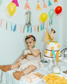 A baby dressed in a costume with a headband and brown booties sits on a table covered with a lace cloth. The table is decorated with snacks, including wafers and chips, and a cake adorned with yellow flowers and a figurine on top. Balloons and colorful decorations hang in the background, creating a festive atmosphere.