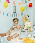 A baby dressed in a costume with a headband and brown booties sits on a table covered with a lace cloth. The table is decorated with snacks, including wafers and chips, and a cake adorned with yellow flowers and a figurine on top. Balloons and colorful decorations hang in the background, creating a festive atmosphere.