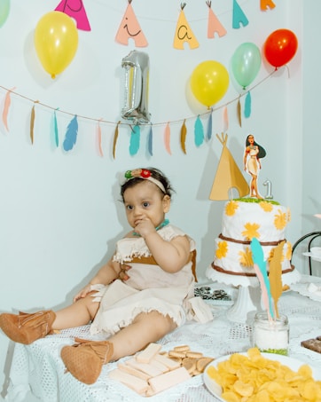 A baby dressed in a costume with a headband and brown booties sits on a table covered with a lace cloth. The table is decorated with snacks, including wafers and chips, and a cake adorned with yellow flowers and a figurine on top. Balloons and colorful decorations hang in the background, creating a festive atmosphere.