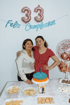 Two people are standing close together at a birthday celebration in front of a white wall with balloons shaped like the number 33 and a 'Feliz Cumpleaños' banner. There is a table in front of them with a cake, plates of chips, cookies, and a photo. Both individuals are smiling and appear happy.