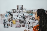 A person is looking at a collage of various photographs and motivational quotes arranged on a white wall. The images and quotes appear to cover a range of activities, people, and scenery, with some text in different languages. The person has long, wavy hair tied up in a ponytail and is wearing an orange and white polka dot shirt.