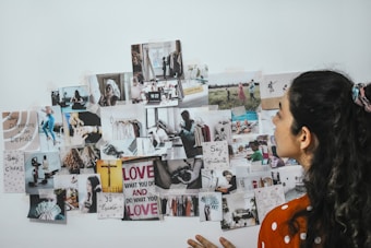 A person is looking at a collage of various photographs and motivational quotes arranged on a white wall. The images and quotes appear to cover a range of activities, people, and scenery, with some text in different languages. The person has long, wavy hair tied up in a ponytail and is wearing an orange and white polka dot shirt.