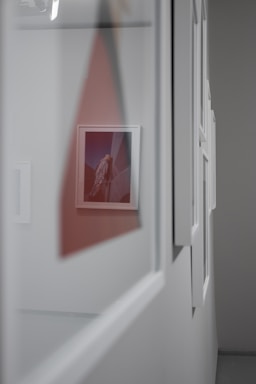 A series of framed pictures are hung on a gray wall, with a perspective that follows the frames into the distance. One central picture is more vividly colored, featuring what appears to be an architectural subject against a sky.