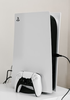 A sleek white gaming console with a modern design, featuring a black logo on its top left. A matching white controller with black accents is placed in front of the console, connected by a cable. The setup is on a plain white surface, creating a minimalist aesthetic.