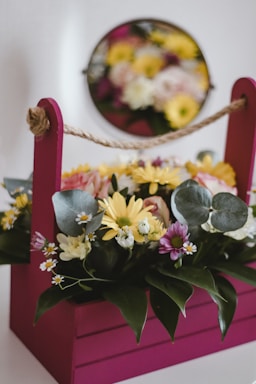 A vibrant floral arrangement sits within a magenta box, featuring a variety of flowers such as yellow daisies, small white blooms, and purple flowers. Green foliage and eucalyptus leaves add texture to the arrangement. In the background, a mirror reflects the flowers, creating a soft, blurred effect.