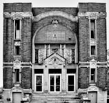 A black-and-white photograph of a historical building with intricate architectural details including brickwork and decorative elements such as stars and menorahs. The facade features large arched windows and a central entrance with glass doors. The building is symmetrical in design.