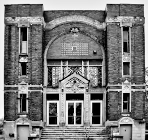 A black-and-white photograph of a historical building with intricate architectural details including brickwork and decorative elements such as stars and menorahs. The facade features large arched windows and a central entrance with glass doors. The building is symmetrical in design.