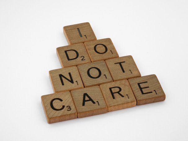 Wooden letter tiles arranged in a pyramid format spell out the phrase 'I DO NOT CARE' on a plain white background. Each tile features a letter with a corresponding numerical value akin to Scrabble tiles.