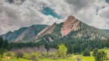 A stunning landscape featuring rugged, angled rock formations known as the Flatirons, rising majestically against a backdrop of a cloudy sky. Lush green trees and grass cover the foreground, providing a stark contrast to the rocky mountains.