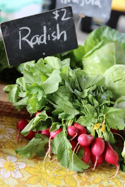 A bunch of fresh radishes with vibrant red bulbs and lush green leaves is placed on a floral-patterned surface. A black sign with the price of $2 is visible, indicating these radishes are for sale. In the background, there are other leafy greens, possibly cabbage or lettuce.