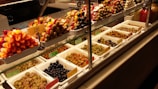 A display case filled with various types of olives and antipasti. The top row features skewers with colorful ingredients like olives, peppers, and cheese. The bottom row consists of neatly arranged trays containing different varieties of olives, each labeled with a price tag.