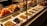 A display case filled with various types of olives and antipasti. The top row features skewers with colorful ingredients like olives, peppers, and cheese. The bottom row consists of neatly arranged trays containing different varieties of olives, each labeled with a price tag.