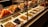 A display case filled with various types of olives and antipasti. The top row features skewers with colorful ingredients like olives, peppers, and cheese. The bottom row consists of neatly arranged trays containing different varieties of olives, each labeled with a price tag.