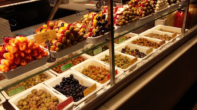 A display case filled with various types of olives and antipasti. The top row features skewers with colorful ingredients like olives, peppers, and cheese. The bottom row consists of neatly arranged trays containing different varieties of olives, each labeled with a price tag.