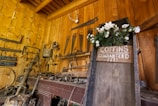 A rustic workshop filled with various antique tools mounted on wooden walls, featuring a prominent wooden sign on a large, weathered plank that reads 'Coffins Guaranteed Fit' adorned with white flowers. The warm tones of the wooden walls and the presence of tools like saws, axes, and hammers create a vintage, historical atmosphere.