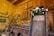 A rustic workshop filled with various antique tools mounted on wooden walls, featuring a prominent wooden sign on a large, weathered plank that reads 'Coffins Guaranteed Fit' adorned with white flowers. The warm tones of the wooden walls and the presence of tools like saws, axes, and hammers create a vintage, historical atmosphere.