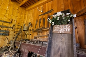 A rustic workshop filled with various antique tools mounted on wooden walls, featuring a prominent wooden sign on a large, weathered plank that reads 'Coffins Guaranteed Fit' adorned with white flowers. The warm tones of the wooden walls and the presence of tools like saws, axes, and hammers create a vintage, historical atmosphere.