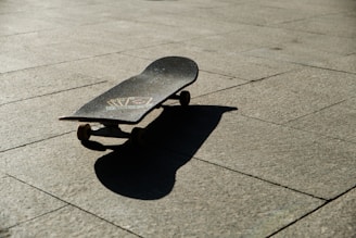 A skateboard lying on a sunlit pavement casting a distinct shadow on the ground. The surface of the skateboard features some graphics, and the surrounding area is clear, emphasizing the texture of the stone pavement.