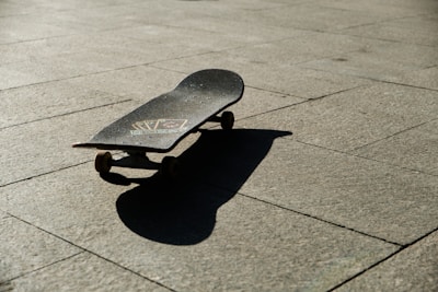 A skateboard lying on a sunlit pavement casting a distinct shadow on the ground. The surface of the skateboard features some graphics, and the surrounding area is clear, emphasizing the texture of the stone pavement.