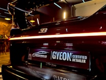 A close-up view of the rear end of a high-performance luxury car with a glossy black finish and a distinctive logo on the back. The car features a sleek tail light that runs across the width of the vehicle. Below the tail light, there is a license plate with a 'GYEON Certified Detailer' logo and another logo that indicates the car has undergone a specialized makeover. The interior setting suggests a high-end detailing shop or garage with warm lighting and modern fixtures in the background.