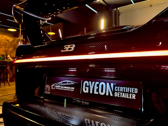 A close-up view of the rear end of a high-performance luxury car with a glossy black finish and a distinctive logo on the back. The car features a sleek tail light that runs across the width of the vehicle. Below the tail light, there is a license plate with a 'GYEON Certified Detailer' logo and another logo that indicates the car has undergone a specialized makeover. The interior setting suggests a high-end detailing shop or garage with warm lighting and modern fixtures in the background.