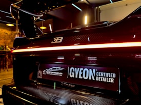 A close-up view of the rear end of a high-performance luxury car with a glossy black finish and a distinctive logo on the back. The car features a sleek tail light that runs across the width of the vehicle. Below the tail light, there is a license plate with a 'GYEON Certified Detailer' logo and another logo that indicates the car has undergone a specialized makeover. The interior setting suggests a high-end detailing shop or garage with warm lighting and modern fixtures in the background.