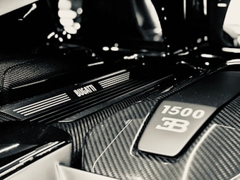 A detailed view of a car engine bay featuring a Bugatti emblem and a label indicating 1500 horsepower. The image highlights the sleek, modern design with prominent carbon fiber elements.