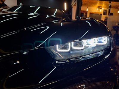 Close-up view of a car's headlight with modern LED lights. The sleek, glossy surface of the car contrasts with the bright white lights that are sharply defined. The reflection of overhead lights is visible on the car's surface, adding a futuristic element.