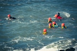 A group of people wearing red helmets and yellow life jackets are swimming together in the ocean. The water is choppy with visible waves, and the individuals appear to be participating in a water-based activity or training exercise.