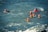 A group of people wearing red helmets and yellow life jackets are swimming together in the ocean. The water is choppy with visible waves, and the individuals appear to be participating in a water-based activity or training exercise.