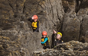 Three individuals wearing helmets and safety gear navigate through a rocky terrain. The person in the front is pointing, possibly indicating a direction or something of interest. The group appears to be engaged in rock climbing or canyoning activities.