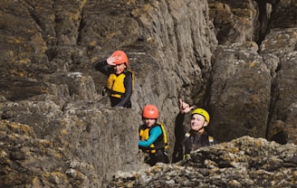 Three individuals wearing helmets and safety gear navigate through a rocky terrain. The person in the front is pointing, possibly indicating a direction or something of interest. The group appears to be engaged in rock climbing or canyoning activities.