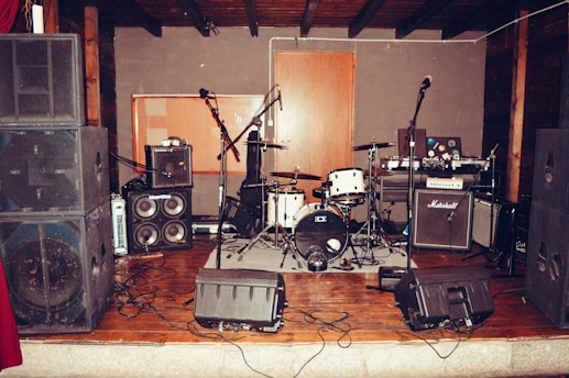 A small music stage is set up with a drum kit at the center, surrounded by various amplifiers and speakers. Microphones stand ready in front of the drums and off to the sides. The wooden floor and ceiling suggest a cozy, intimate venue. Cables are strewn across the floor, leading to the equipment.