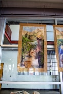 A large fabric or canvas banner depicting a pastoral scene hangs in front of a glass wall. The artwork shows a person wearing a hat, likely engaged in agricultural activities, amidst rows of plants in a field with mountains in the background. Baskets are filled with gathered crops at the person's feet.