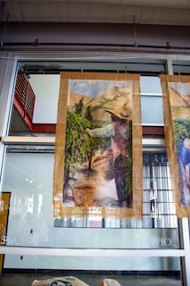 A large fabric or canvas banner depicting a pastoral scene hangs in front of a glass wall. The artwork shows a person wearing a hat, likely engaged in agricultural activities, amidst rows of plants in a field with mountains in the background. Baskets are filled with gathered crops at the person's feet.