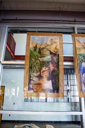 A large fabric or canvas banner depicting a pastoral scene hangs in front of a glass wall. The artwork shows a person wearing a hat, likely engaged in agricultural activities, amidst rows of plants in a field with mountains in the background. Baskets are filled with gathered crops at the person's feet.