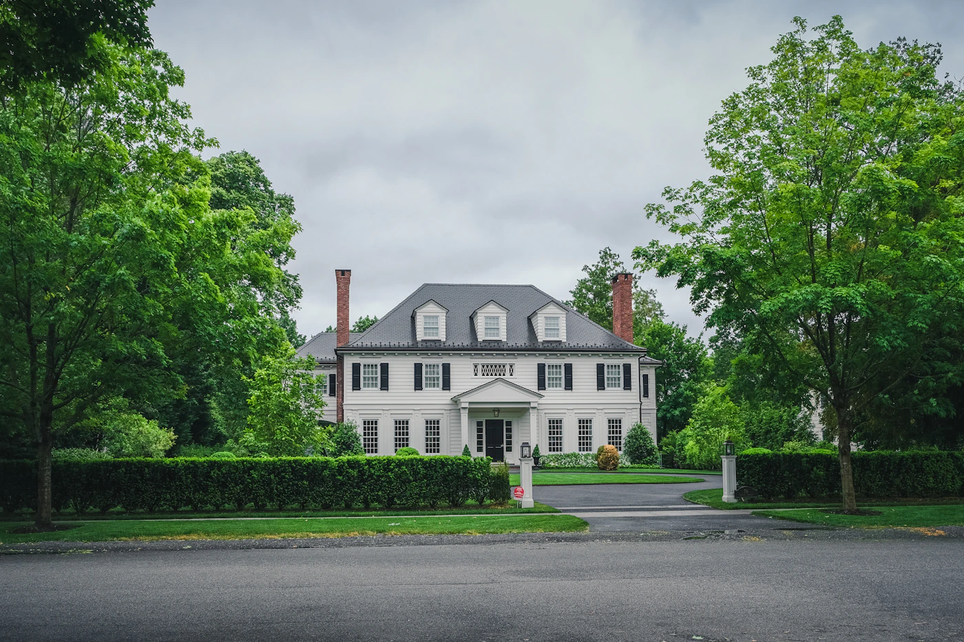 A Georgian-style white manor house with black shutters, twin chimneys, and a tree-lined driveway, set in green grounds under an overcast sky