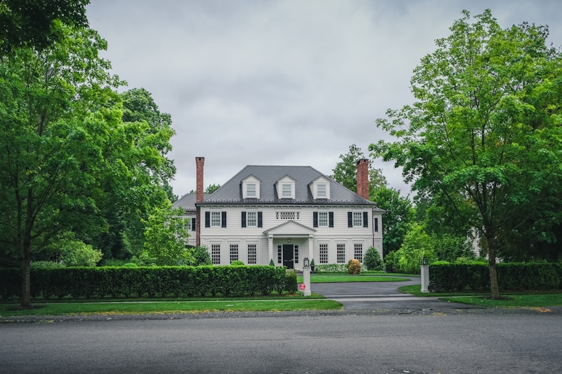A large, elegant white colonial-style house is situated in the center, surrounded by lush green trees and shrubs. The house has a symmetrical design with multiple windows, a prominent central entrance, and two brick chimneys. The grey roof complements the serene and well-maintained garden landscape.