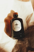 A dark glass bottle with a wooden cap is displayed on a textured, cream-colored fabric. The label on the bottle reads 'Brut Mojito' and features ornate typography and design. Warm lighting and soft shadows create a cozy and artisanal ambiance.