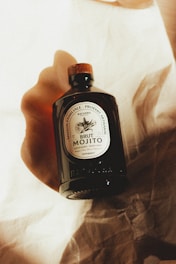 A dark glass bottle with a wooden cap is displayed on a textured, cream-colored fabric. The label on the bottle reads 'Brut Mojito' and features ornate typography and design. Warm lighting and soft shadows create a cozy and artisanal ambiance.