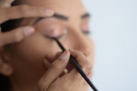 A close-up view of a person applying eyeliner to their eyelid with a black brush. The hand is steady, focusing on precision, while the eyelid is gently held taut.