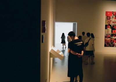 A dimly lit gallery space with several people viewing exhibits. On the left, a person in a dark shirt is interacting with a digital display mounted on the wall. Towards the back, more people are seen walking and standing near a bright doorway, and on the right, there is a wall with a colorful collage of images.