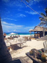 A pristine beach setting with white sand and clear blue skies. There are wooden beach chairs and tables scattered around, some under large umbrellas. A tiki-style bar with a thatched roof is visible to the right, and string lights hang above. Palm trees frame the scene, and the ocean gently laps the shore in the background. Several people are leisurely enjoying the beach atmosphere.