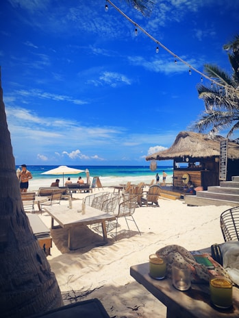A pristine beach setting with white sand and clear blue skies. There are wooden beach chairs and tables scattered around, some under large umbrellas. A tiki-style bar with a thatched roof is visible to the right, and string lights hang above. Palm trees frame the scene, and the ocean gently laps the shore in the background. Several people are leisurely enjoying the beach atmosphere.