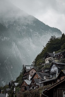 A picturesque mountainside village with a steep incline covered in lush green trees. Traditional wooden and stone houses with dark roofs are nestled closely together, creating a quaint and rustic atmosphere. The background features towering mountains shrouded in mist, adding a mysterious and serene feel to the scene.