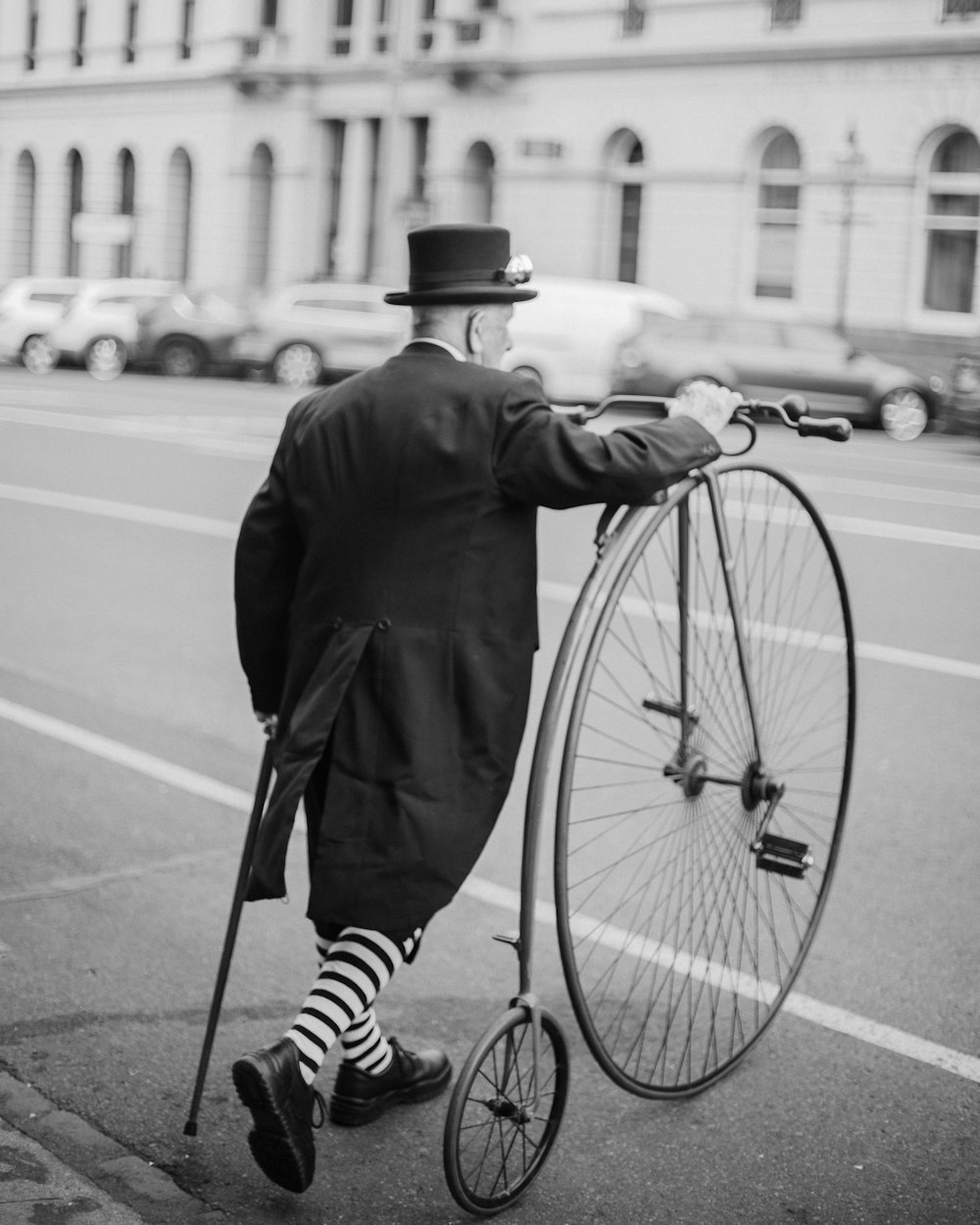 uomo in cappotto nero e pantaloni in bicicletta sulla strada in fotografia in scala di grigi